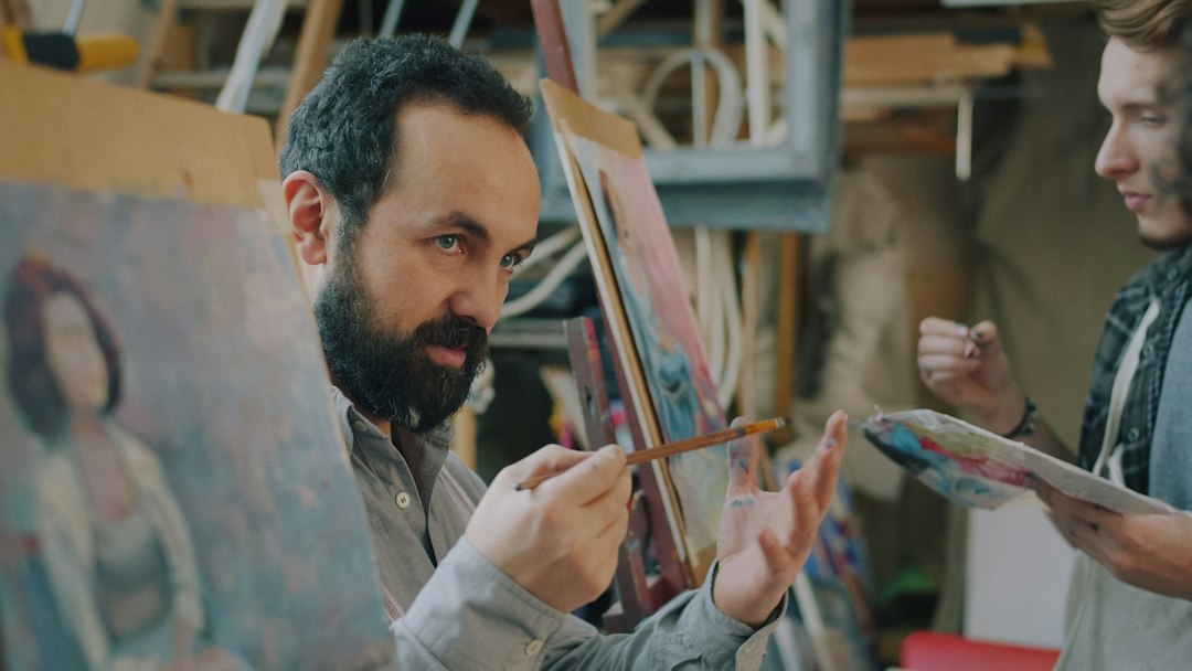 A bearded man holding a paintbrush in an art studio with another person
