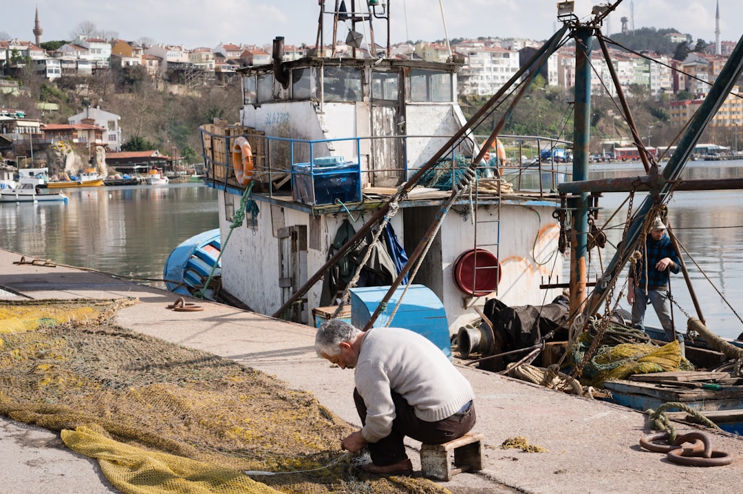 Elderly man repairs fishing nets by a docked boat.